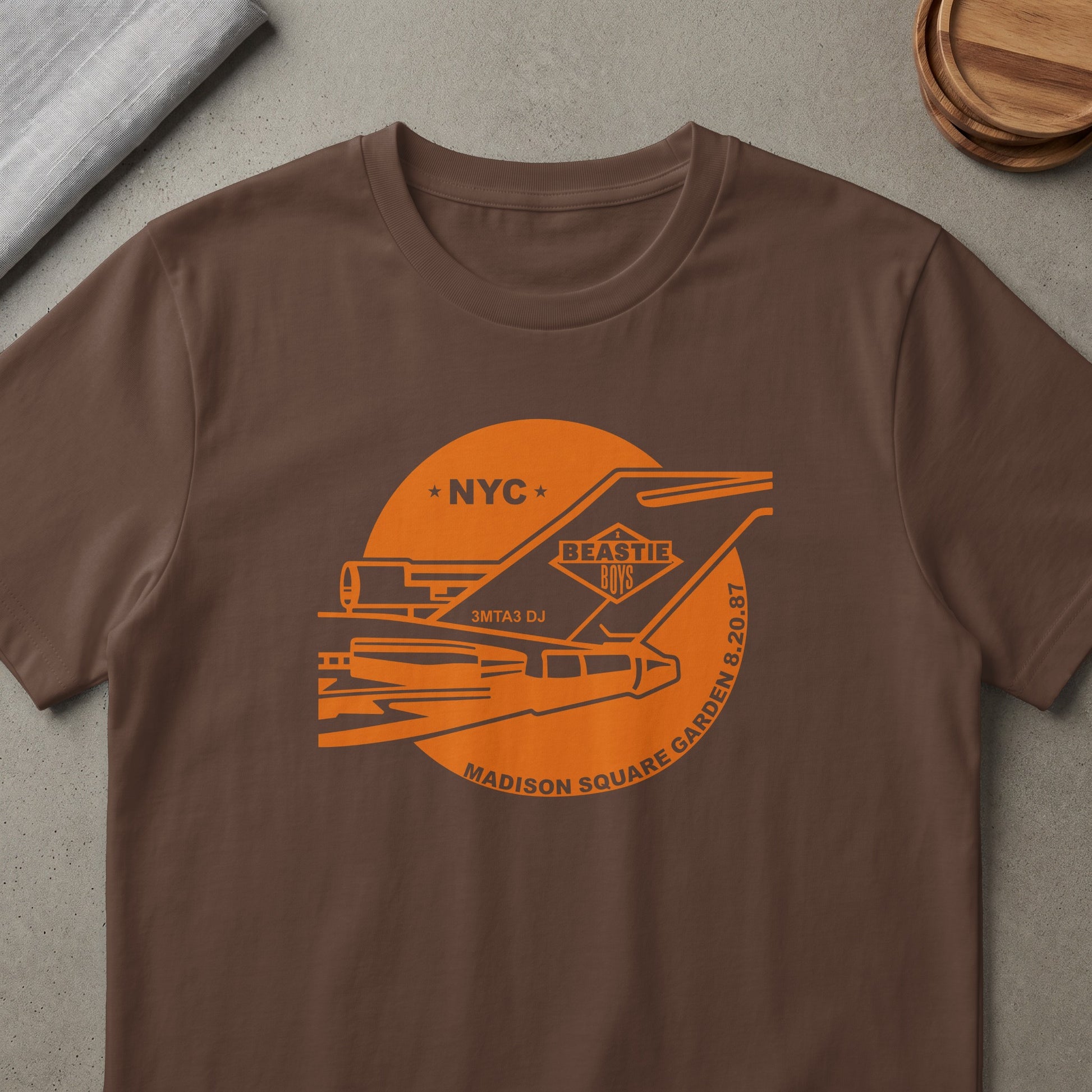 Brown t-shirt with orange Beastie Boys graphic featuring a plane, NYC, and Madison Square Garden 8.20.87.